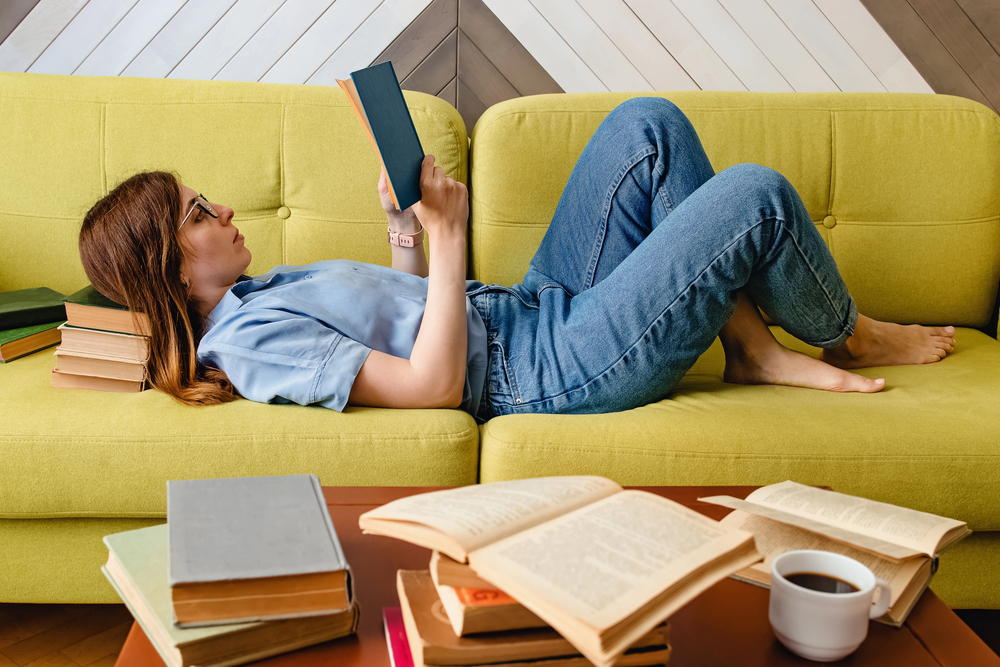 Frau liest entspannt auf einem grünen Sofa, umgeben von mehreren Büchern
