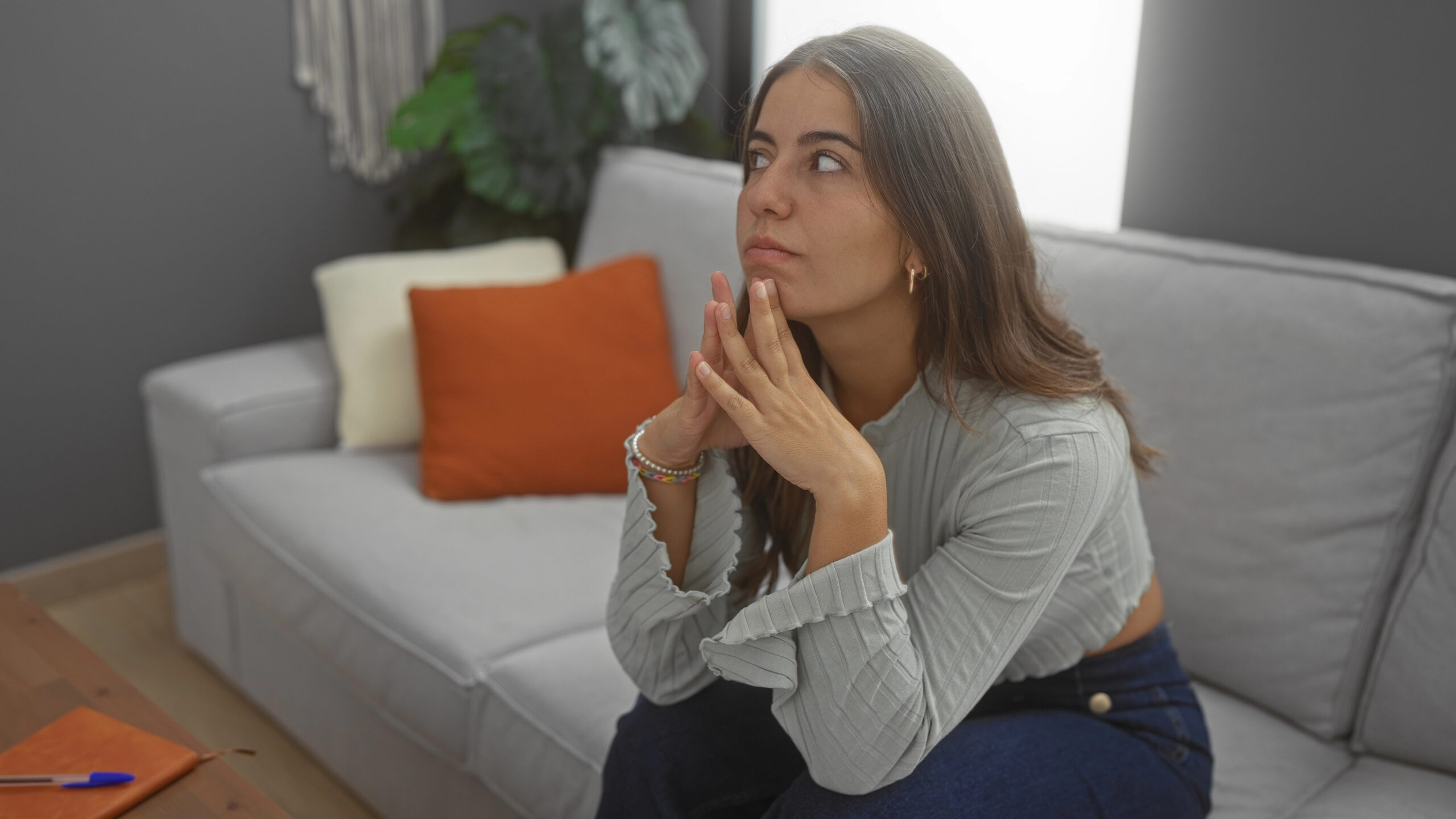 Eine junge Frau mit langen braunen Haaren sitzt vor einem Sofa, die Hände gefaltet vor dem Gesicht, und blickt nachdenklich zur Seite – ein Bild für das Nachgrübeln und die innere Gedankenspirale nach einem Gespräch