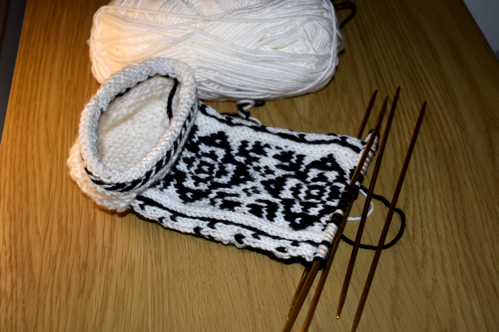 Angefangene Strickarbeit mit schwarz-weißem Muster auf Nadeln, daneben ein Knäuel weißer Wolle auf einem Holztisch