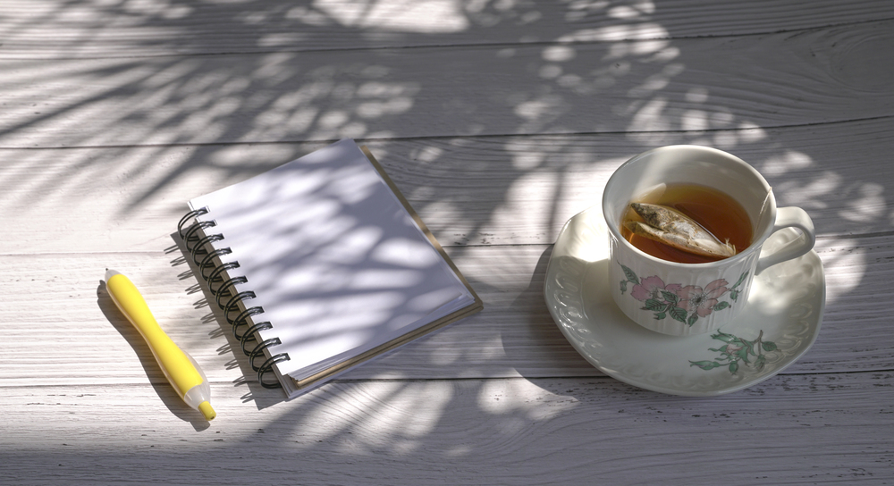 Spiralnotizbuch, gelber Stift und eine Tasse Tee auf einem hellen Holztisch im Licht- und Schattenspiel von Blättern