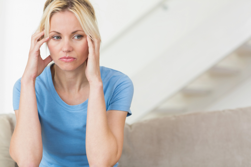 Blonde Frau im blauen T-Shirt hält sich die Hände an die Schläfen und wirkt nachdenklich – Symbolbild für innere Anspannung und Stimmungsschwankungen.
