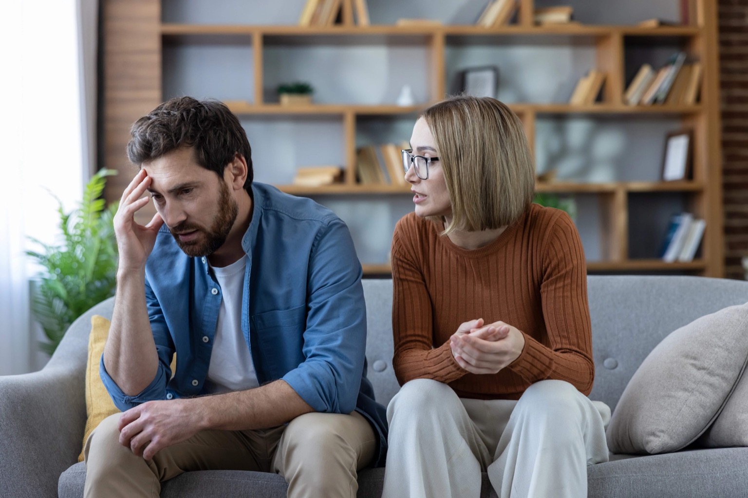 Ein Paar sitzt auf einem Sofa in angespannter Gesprächssituation; der Mann wirkt belastet, während die Frau sich ihm zuwendet.