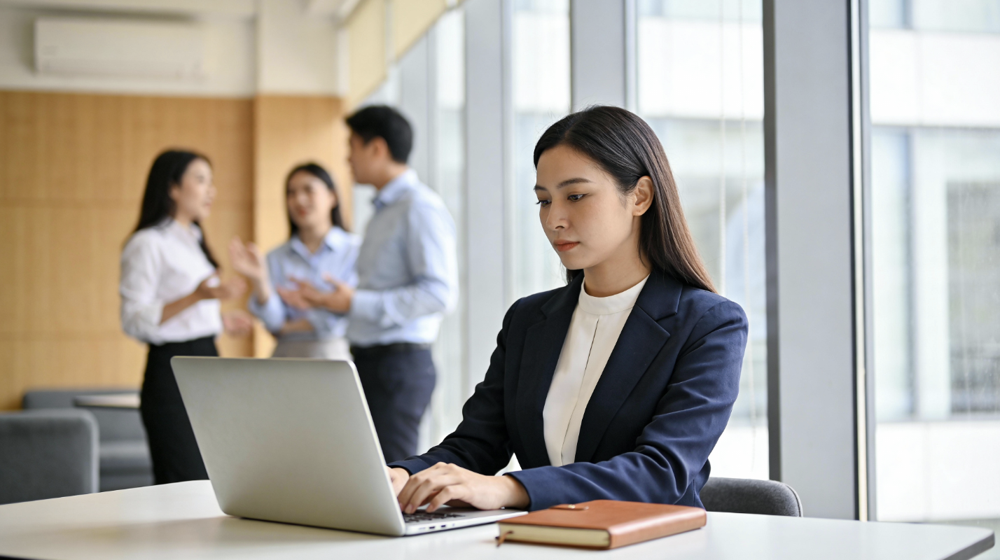 Eine Frau im Business-Outfit sitzt konzentriert an einem Laptop an einem Tisch, während im Hintergrund drei Kolleg:innen im Stehen miteinander sprechen. Sie wirkt ruhig und etwas abseits vom Gruppengespräch.
