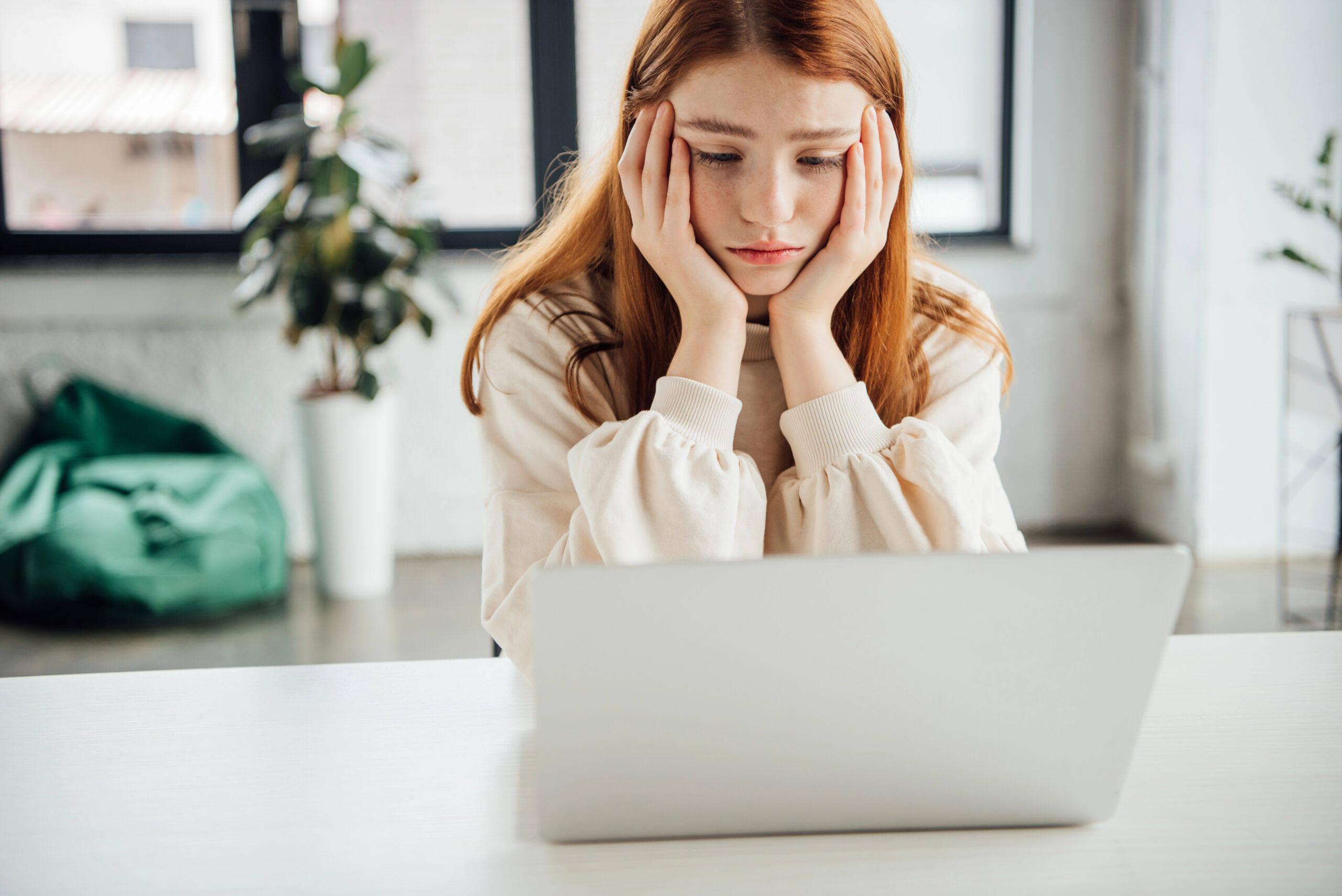 Junge Frau mit roten Haaren sitzt vor einem Laptop, stützt den Kopf in die Hände und blickt nachdenklich und bedrückt auf den Bildschirm – sie wirkt, als würde sie sich Sorgen machen oder sich Vorwürfe machen.