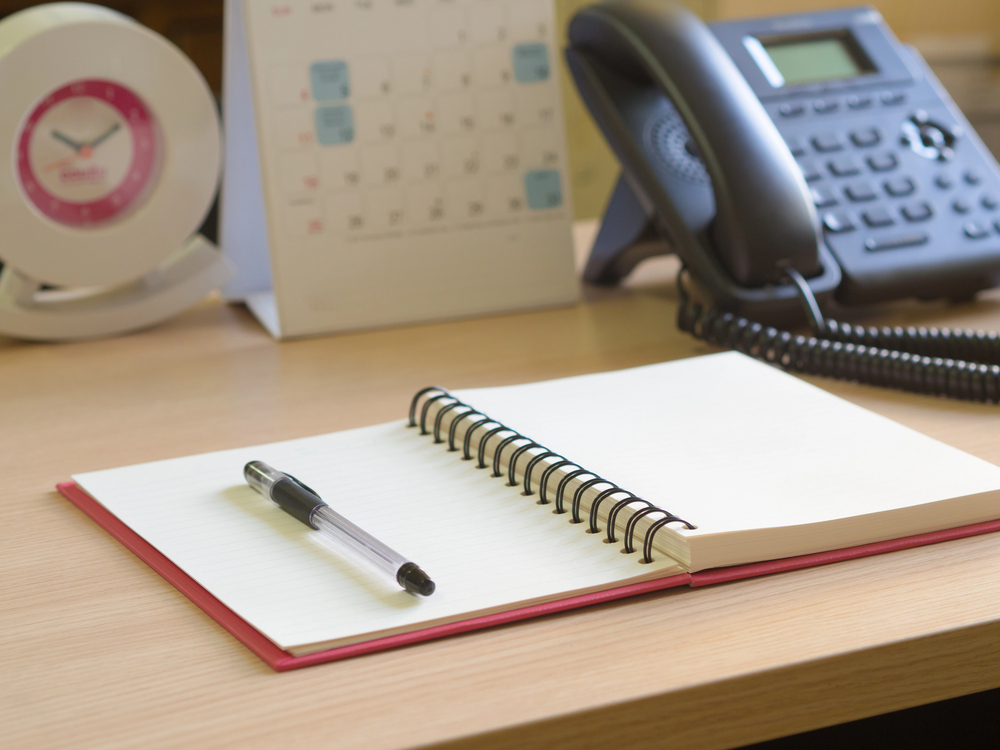 Aufgeschlagenes Notizbuch mit Kugelschreiber auf einem Schreibtisch, dahinter ein Telefon, ein Tischkalender und eine Uhr