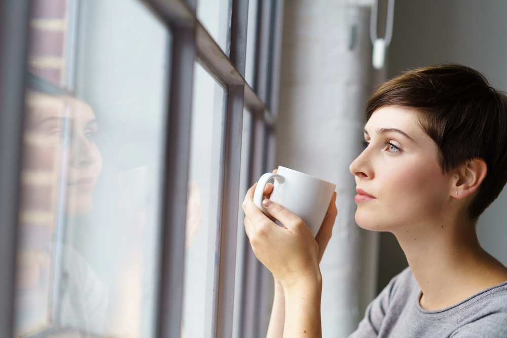 Eine Frau mit kurzen braunen Haaren hält eine weiße Tasse in beiden Händen und schaut aus dem Fenster; ihr Gesicht spiegelt sich in der Scheibe.