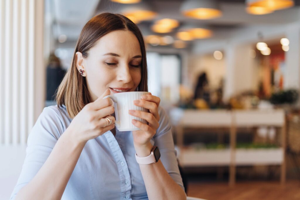 Eine Frau mit geschlossenen Augen hält eine Tasse in beiden Händen und lächelt sanft. Sie sitzt in einem gemütlichen Café und genießt einen ruhigen Moment.