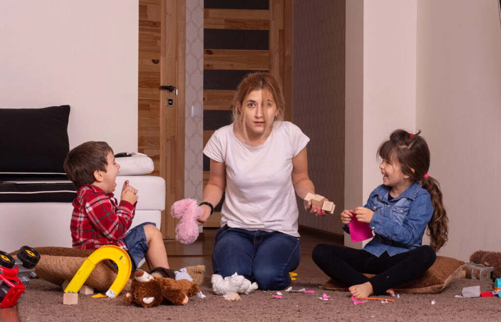 Wohnzimmer: Eine erschöpfte Bezugsperson sitzt am Boden zwischen zwei spielenden Kindern; Spielzeug liegt herum – Stimmung: laut, trubelig.