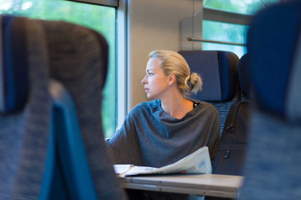 Person sitzt im Zug am Fenster, schaut nach draußen; auf dem Tisch liegt eine Zeitung – ruhiger Moment während der Fahrt.