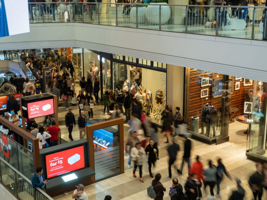 Blick auf eine belebte Shopping-Mall mit vielen Menschen auf mehreren Ebenen, die an Geschäften vorbeigehen und einkaufen.