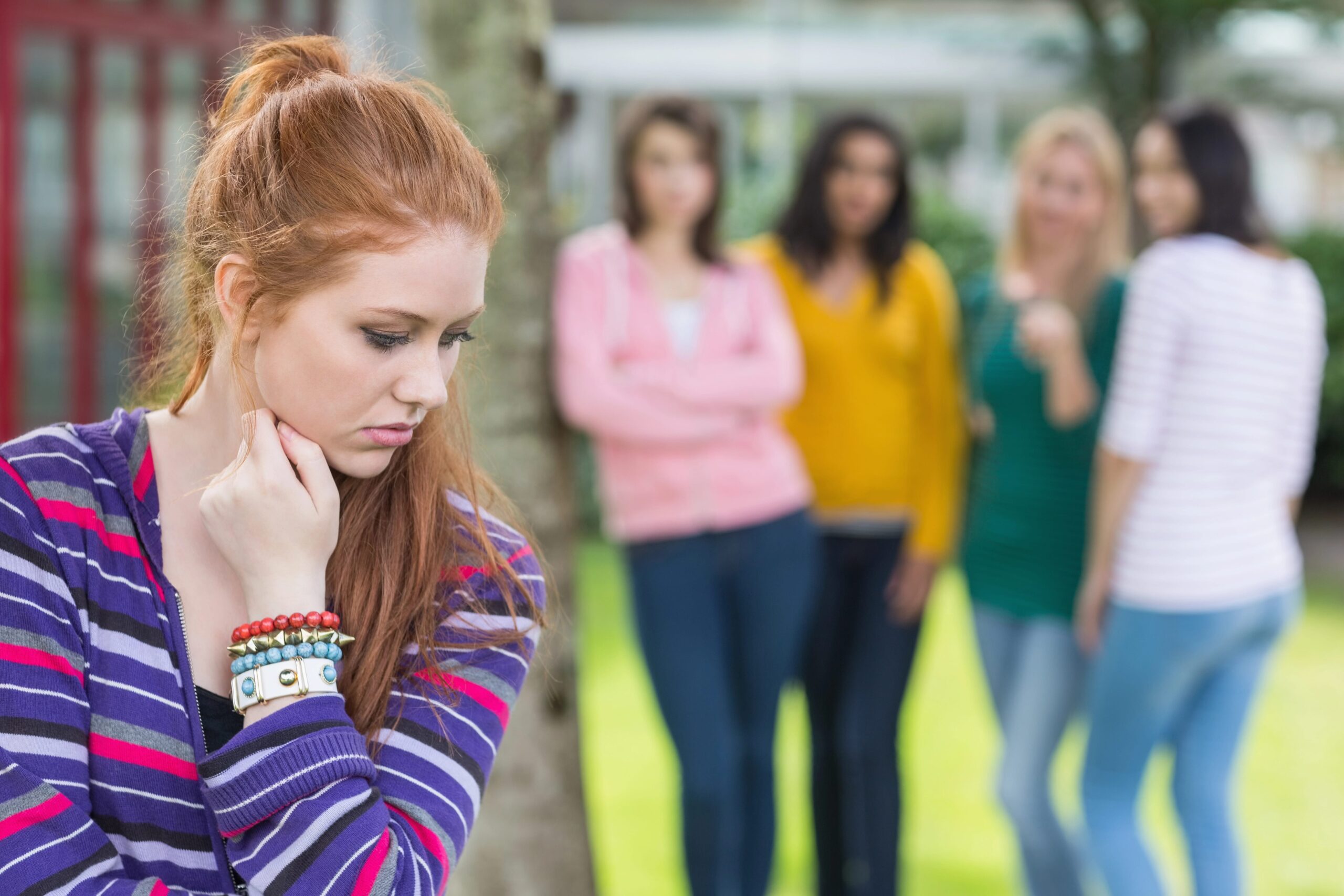 Junge Frau steht alleine und nachdenklich im Vordergrund, während sie von einer Gruppe lachender Freundinnen im Hintergrund beobachtet wird.