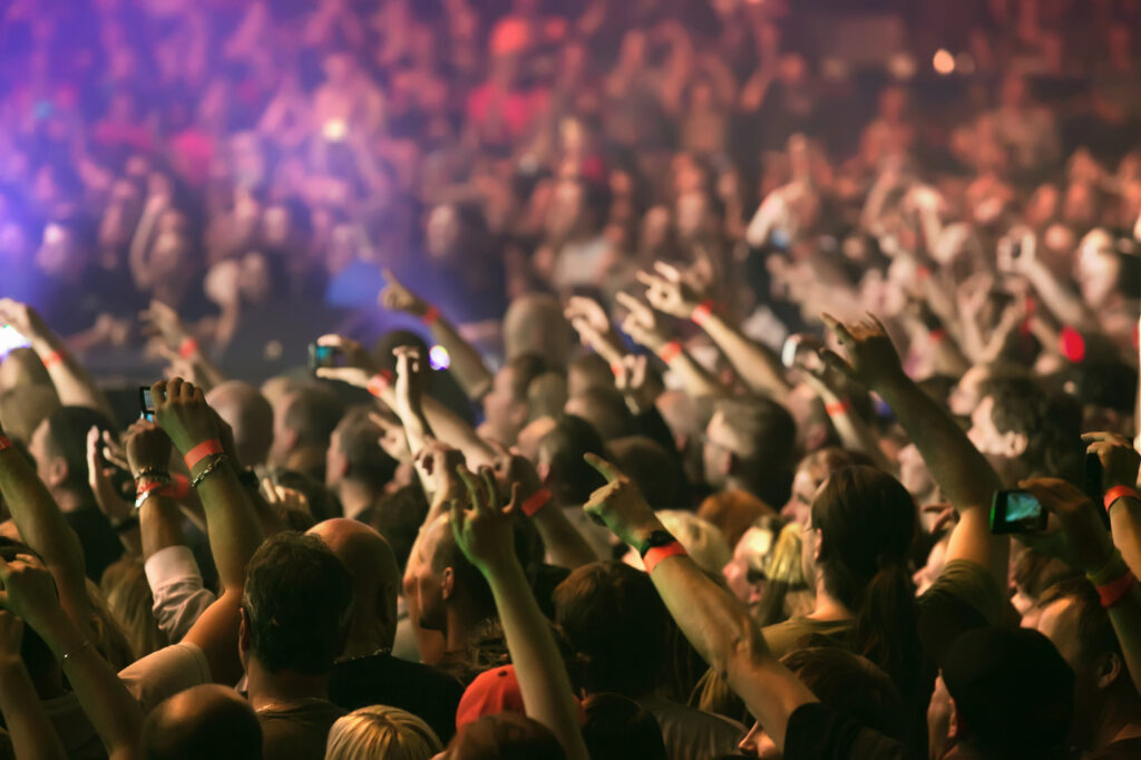 Große Menschenmenge auf einem Konzert, viele Arme in der Luft und bunte Beleuchtung im Hintergrund.