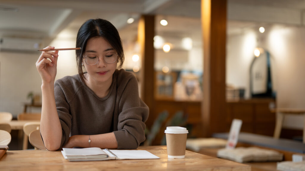 Eine junge Frau sitzt an einem Tisch, schaut nachdenklich auf ein Notizbuch, hält einen Stift in der Hand und ist von einer Tasse Kaffee umgeben – symbolisiert das Schreiben eines Interessen-Tagebuchs.