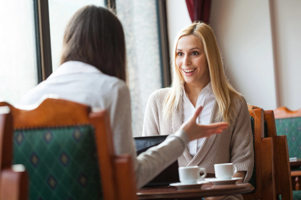 Eine blonde Frau mit einem begeisterten Lächeln führt ein angeregtes Gespräch in einem Café.