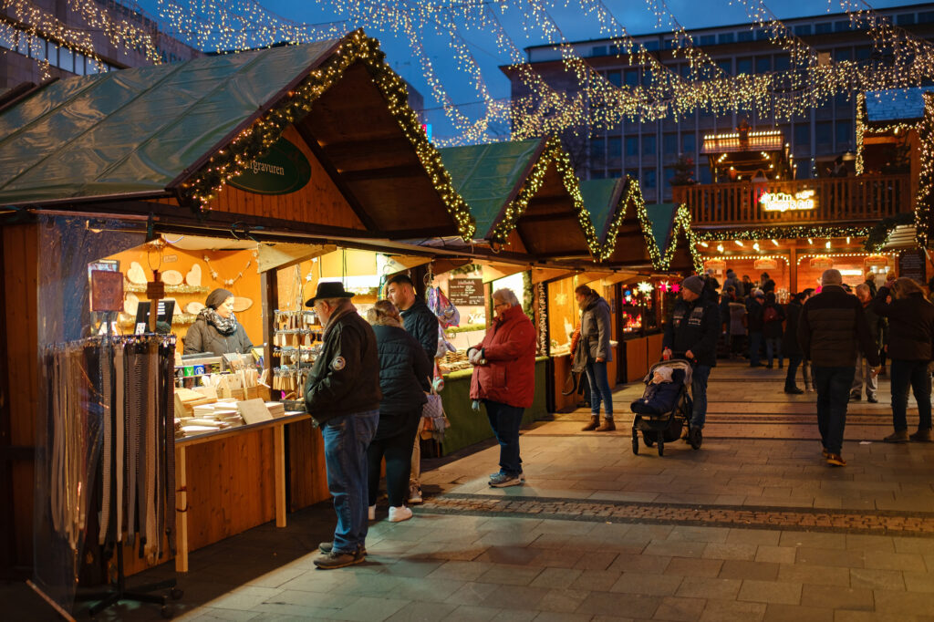 Menschen bummeln an festlich beleuchteten Holzständen eines Weihnachtsmarktes entlang.