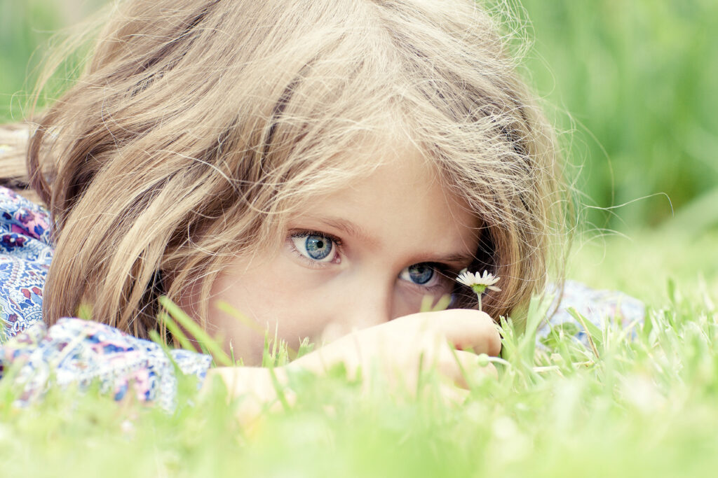 Ein kleines Mädchen mit blauen Augen liegt im Gras und betrachtet eine Gänseblümchenblüte, die sie in der Hand hält.