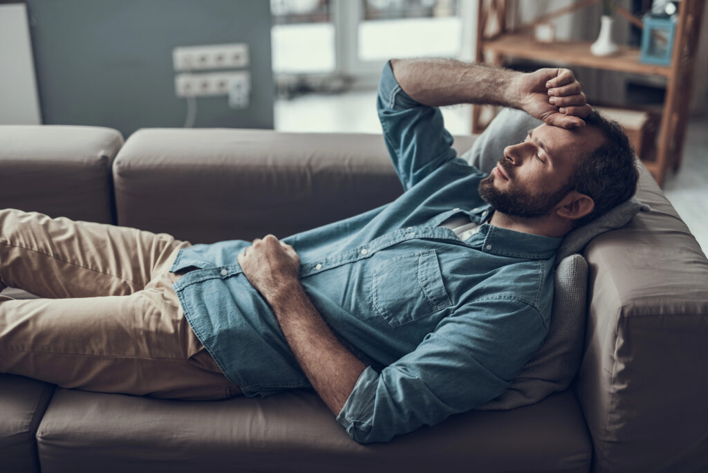 Erschöpfter Mann liegt auf dem Sofa und hält sich die Hand vor die Stirn