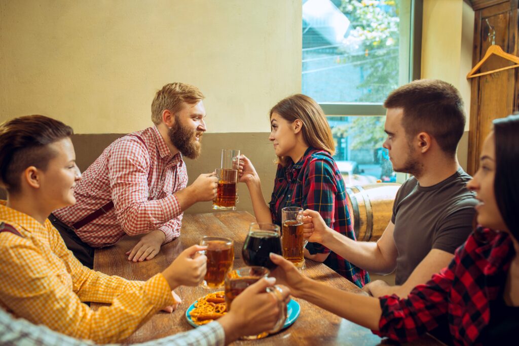 Gruppe von Freunden genießt Getränke in einer Bar, während ein Mann skeptisch das Gespräch zwischen einem anderen Mann und einer Frau beobachtet.