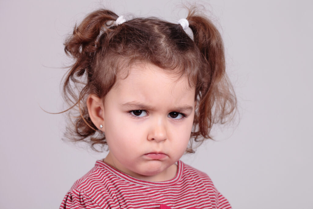 Kleines Mädchen mit mürrischem Gesichtsausdruck und Zöpfen steht vor einem hellgrauen Hintergrund.
